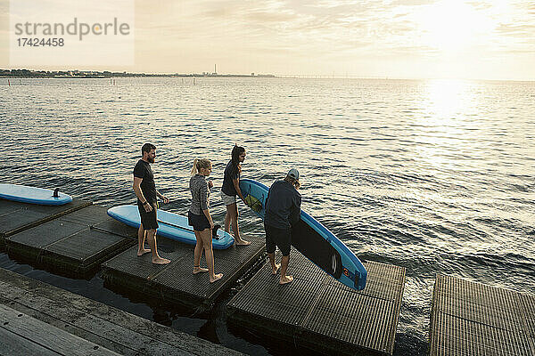 Männliche und weibliche Freunde tragen ein Paddelbrett  während sie auf einem Pier am Meer stehen