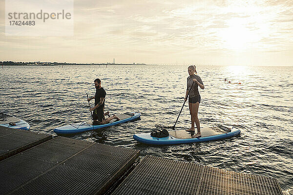 Männliche und weibliche Freunde rudern Paddleboard durch Pier im Meer