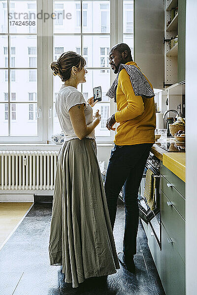 Lächelnde Frau mit Smartphone im Gespräch mit ihrem Freund bei der Hausarbeit in der Küche in voller Länge