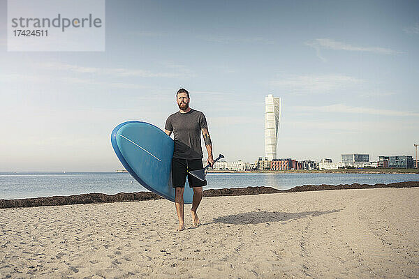 Mann geht mit Stand Up Paddleboard gegen den Himmel am Strand