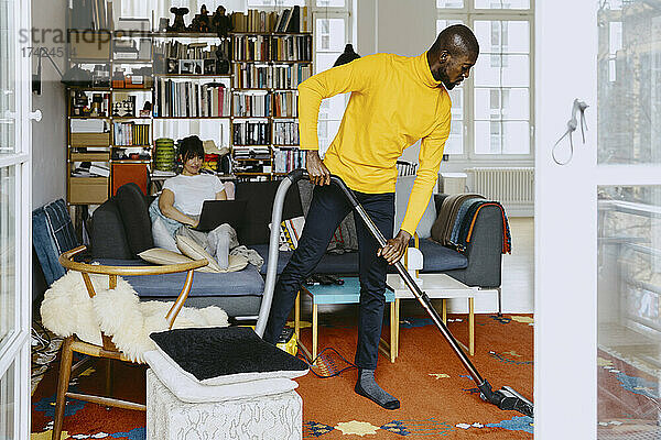 Mann reinigt Teppich in voller Länge mit Staubsauger  während eine Freiberuflerin im Wohnzimmer zu Hause am Laptop arbeitet
