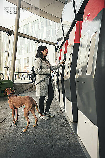 Junge Frau benutzt einen Fahrkartenautomaten  während sie mit ihrem Hund am Bahnhof pendelt