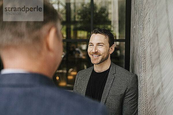Lächelnder Geschäftsmann  der einen männlichen Fachmann ansieht  der während eines Seminars im Kongresszentrum an der Wand steht und diskutiert