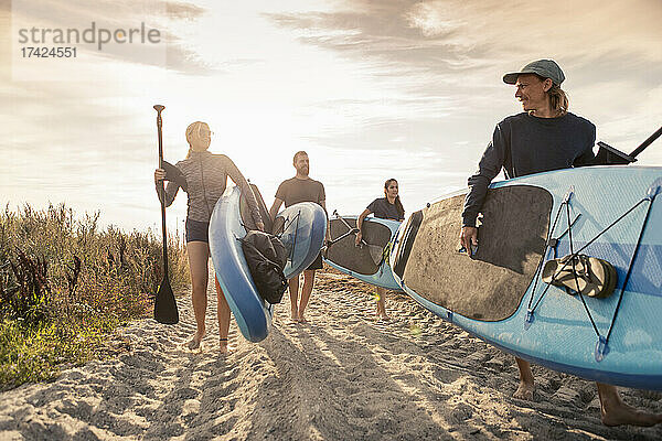 Männliche und weibliche Freunde tragen Paddleboard am Sandstrand bei Sonnenuntergang