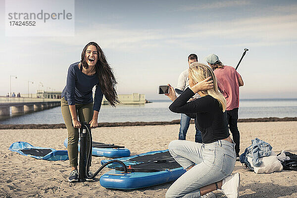 Glückliche Frau  die ein Paddleboard aufpumpt  während eine Freundin am Strand fotografiert