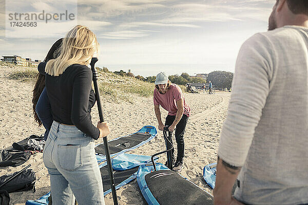 Mann pumpt Stand Up Paddleboard  während Freunde am Strand stehen