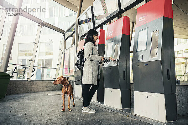 Seitenansicht einer Frau mit Hund an einem Fahrkartenautomaten im Bahnhof