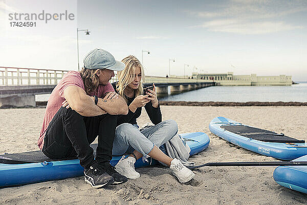 Frau  die ein Smartphone benutzt  während ein männlicher Freund auf einem Paddelbrett sitzt