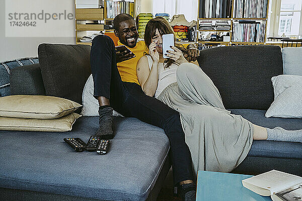 Paar in voller Länge  das lacht  während es ein Smartphone benutzt und zusammen auf dem Sofa im Wohnzimmer sitzt