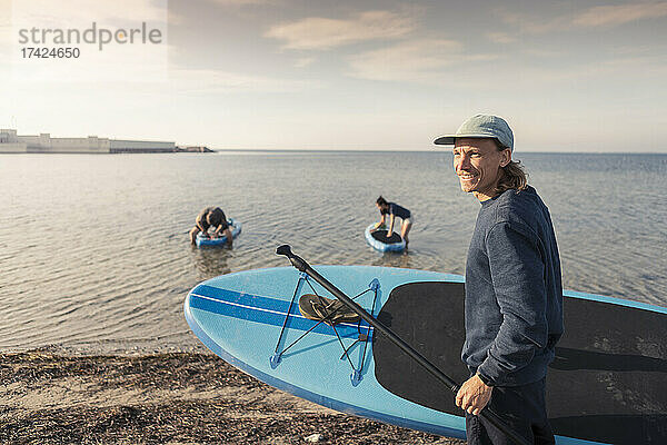 Männlicher Lehrer mit Paddleboard am Strand bei Sonnenuntergang