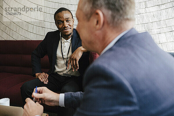 Lächelnder Geschäftsmann  der einen männlichen Kollegen ansieht  während er auf dem Sofa bei einer Networking-Veranstaltung sitzt und diskutiert