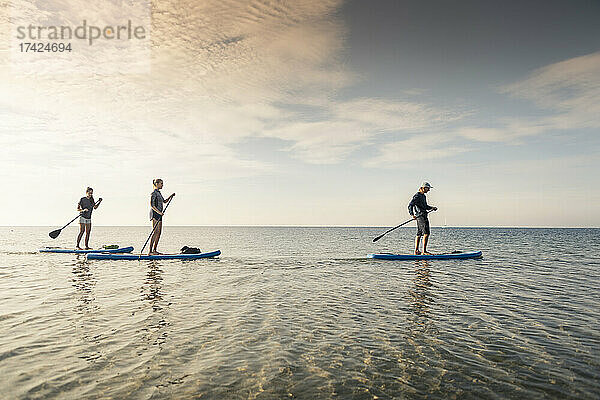 Weibliche Freunde lernen Paddleboarding von einem männlichen Lehrer im Meer bei Sonnenuntergang