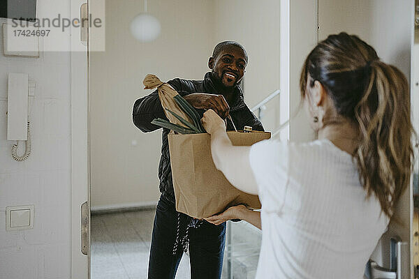 Ein lächelnder Mann gibt einer an der Tür stehenden Frau eine Papiertüte mit Lebensmitteln