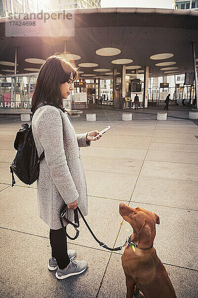 Seitenansicht einer Frau  die ihr Smartphone benutzt  während sie mit ihrem Hund auf dem Gehweg steht