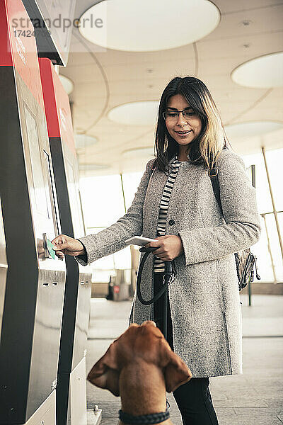 Frau schaut Hund an  während sie am Bahnhof eine Fahrkarte kauft