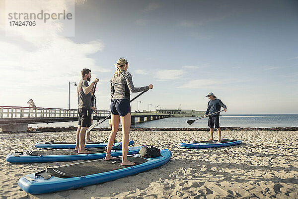 Männlicher Lehrer unterrichtet Mann und Frau am Strand im Paddleboarding