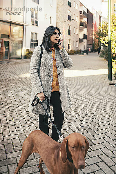 Junge Frau spricht in ihr Smartphone  während sie mit ihrem Hund auf dem Gehweg spazieren geht