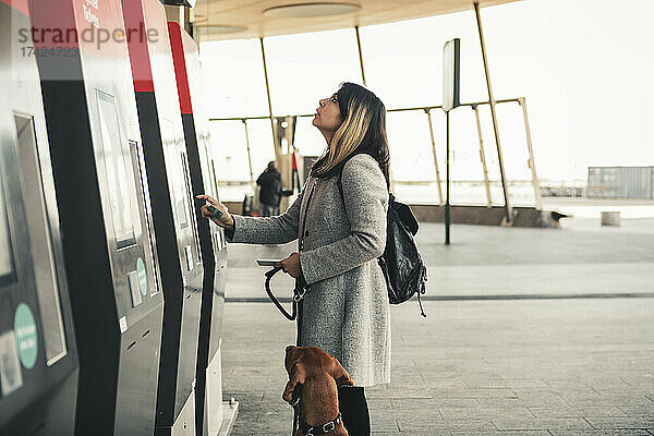 Junge Frau mit Hund zahlt mit Kreditkarte beim Kauf einer Fahrkarte am Bahnhof