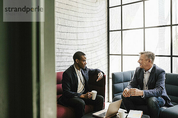 Männlicher Unternehmer im Gespräch mit einem reifen Geschäftsmann  der auf einem Sofa im Kongresszentrum sitzt