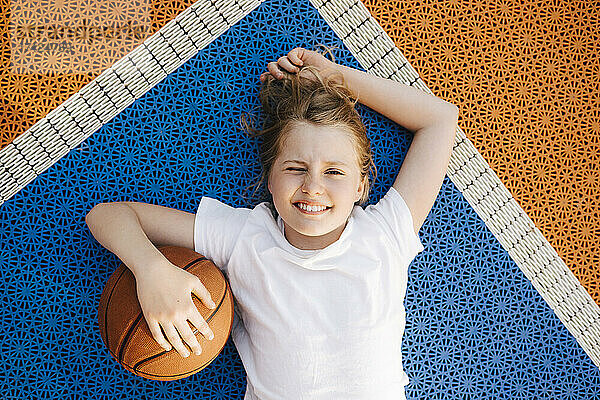 Vorpubertäres Mädchen mit Basketball  das mit den Augen zwinkert  während es auf dem Spielfeld liegt