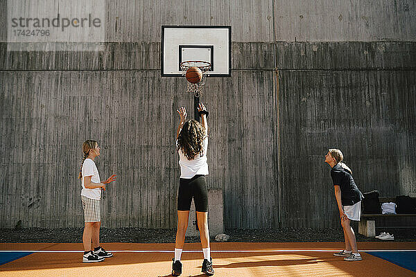 Rückansicht eines Mädchens  das beim Spielen mit Freunden auf einem Sportplatz einen Basketball in einen Korb wirft