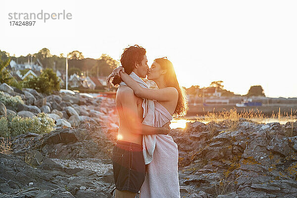 Junges Paar  das sich küsst  während es bei Sonnenuntergang auf einem Felsen am Meer steht