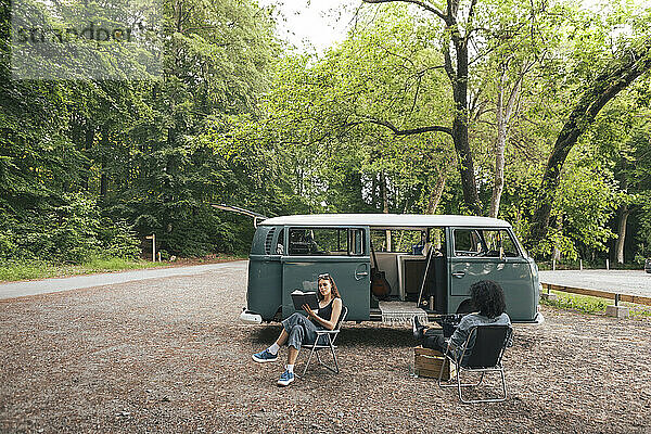 Rückansicht eines Mannes  der einen Laptop benutzt  während seine Freundin vor einem Campingwagen ein Buch liest