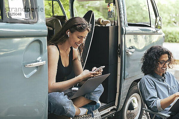 Lächelnde Frau  die ein Smartphone benutzt  während ein männlicher Freund mit einem Buch vor einem Campingwagen sitzt