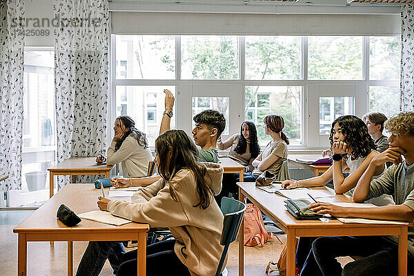 Junge hebt die Hand  während er mit anderen Schülern im Klassenzimmer sitzt