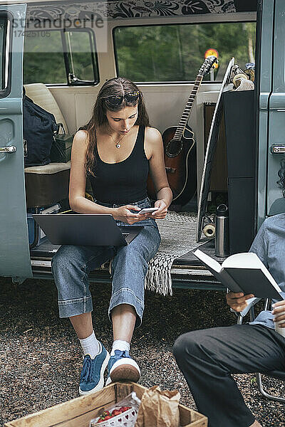 Frau benutzt Smartphone  während männlicher Freund mit Buch im Urlaub sitzt