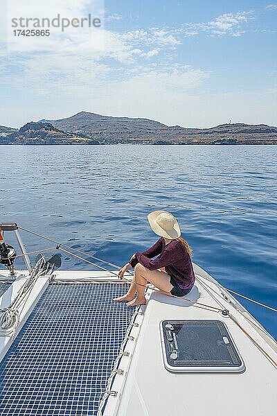 Junge Frau mit Hut sitzt auf Deck am Netz eines Segel-Katamaran  Segeltörn  Rhodos  Dodekanes  Griechenland  Europa