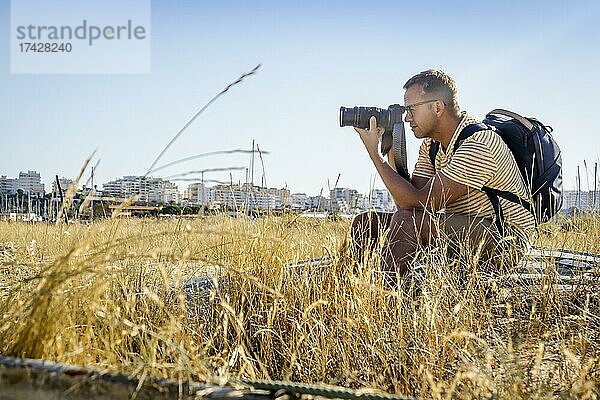 Ein Fotograf mit einem Rucksack sitzt im trockenen Gras und richtet seine Kamera nach vorne  mit der Stadt im Hintergrund