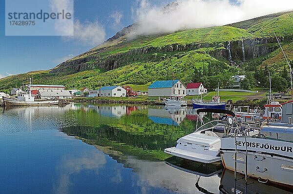 Boote und kleine Häuser spiegeln sich im Fjord  Wasserfall an grünem Berghang  Seydisfjördur  Island  Europa