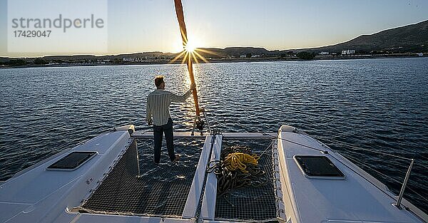 Junger Mann steht am Vorsegel  Sonnenuntergang mit Sonnenstern  Beim Segeln mit einem Segel-Katamaran  Dodekanes  Griechenland  Europa