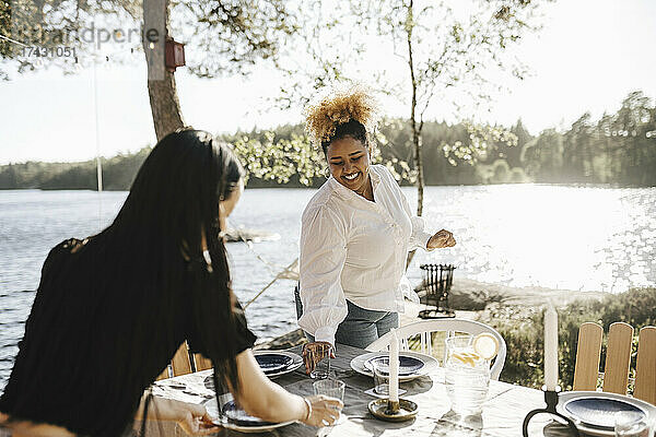 Lächelnde Frau deckt den Tisch mit ihrer Freundin während einer Dinnerparty an einem sonnigen Tag