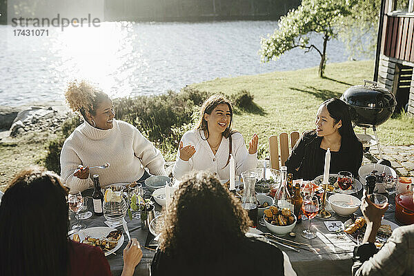 Glückliche Frau im Gespräch mit weiblichen Freunden  während sie eine Dinnerparty im Hinterhof an einem sonnigen Tag genießt