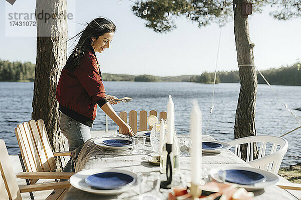 Lächelnde junge Frau deckt den Tisch am See an einem sonnigen Tag