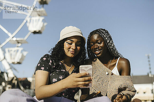 Lächelnde Freundinnen  die ein Smartphone gegen den klaren Himmel an einem sonnigen Tag benutzen