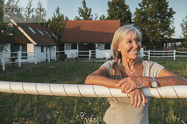 Lächelnde ältere Landarbeiterin lehnt am Zaun eines Bauernhofs