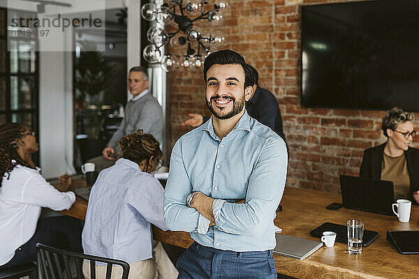 Porträt eines lächelnden männlichen Unternehmers  der mit verschränkten Armen dasteht  während sich im Hintergrund Kollegen unterhalten