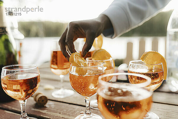 Abgeschnittene Hand einer Frau  die eine Orangenscheibe auf einem Weinglas am Tisch hält