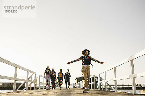 Teenager-Mädchen auf dem Skateboard  während ihre Freunde auf dem Pier gegen den klaren Himmel laufen