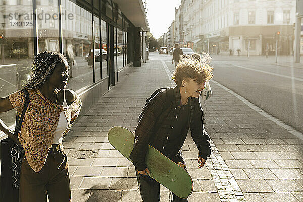 Jugendlicher mit Skateboard  der lachend neben seiner Freundin auf einem Fußweg in der Stadt steht