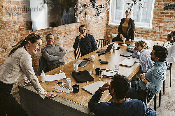 Reife Geschäftsfrau  die sich auf einen Konferenztisch stützt  während sie mit Kollegen im Sitzungssaal einen Plan bespricht