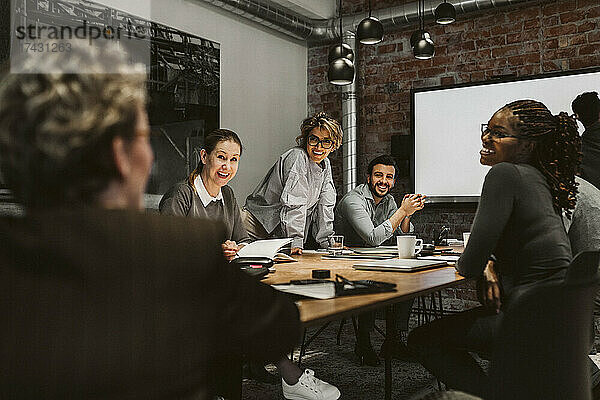 Lächelnde männliche und weibliche Fachleute  die einen reifen Unternehmer ansehen  während sie am Konferenztisch sitzen