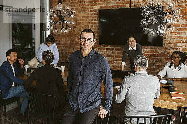 Porträt eines lächelnden Geschäftsmannes im Stehen  während Kollegen im Hintergrund diskutieren