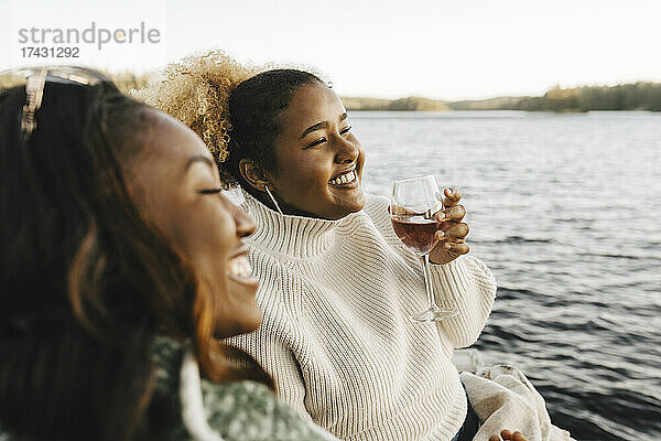 Lächelnde Frau genießt Wein mit Freundin am Seeufer
