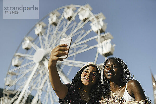 Multirassische Freundinnen machen ein Selfie mit ihrem Smartphone vor einem Riesenrad an einem sonnigen Tag