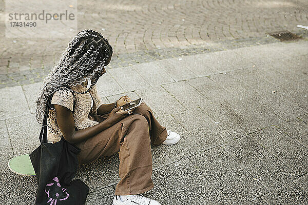 Junge Frau  die ein Mobiltelefon benutzt  während sie auf dem Gehweg sitzt