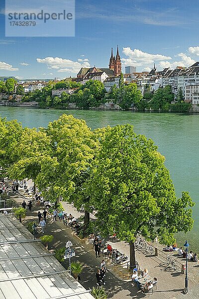 Blick von der Flusspromenade auf das Basler Münster und die Basler Altstadt mit dem türkis farbigem Rhein Fluss und der belebten Uferpromenade im Vordergrund